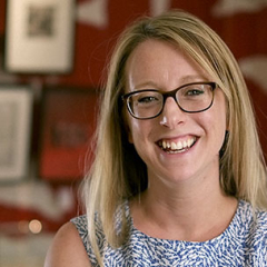 Portrait of a woman with blonde hair, wearing glasses and a blue patterned dress, smiling in front of a red background.