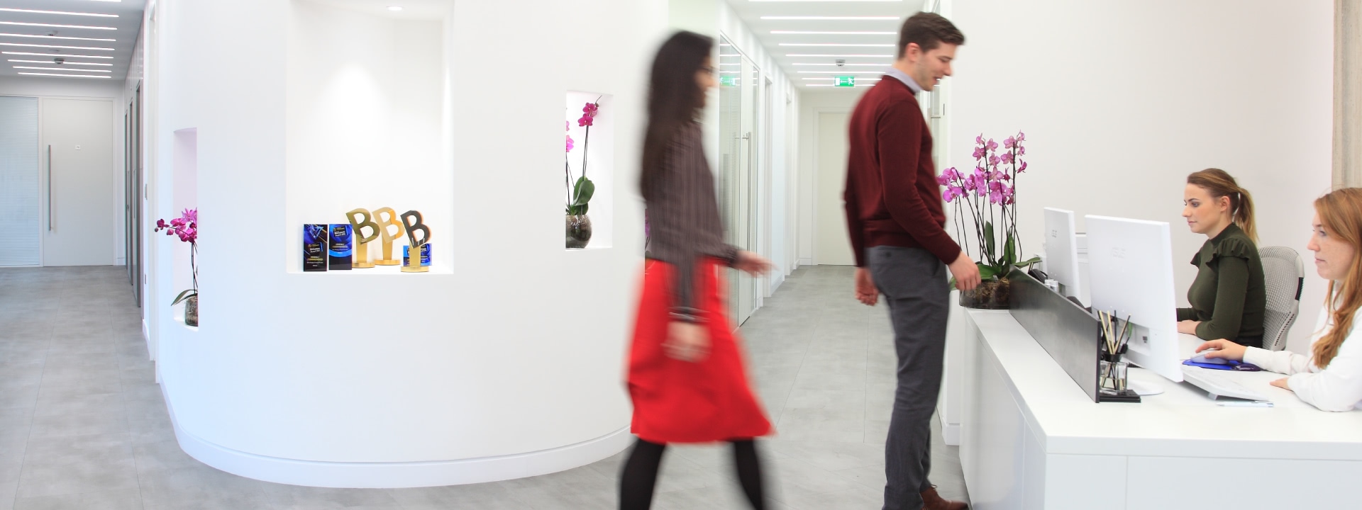 Modern office reception with two people. A woman in a red skirt walks past a man at the reception desk. White walls feature decorative niches with awards and pink orchids. Visible text includes 