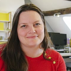 Portrait of a woman with long brown hair, wearing a red sweater, smiling in an office setting.