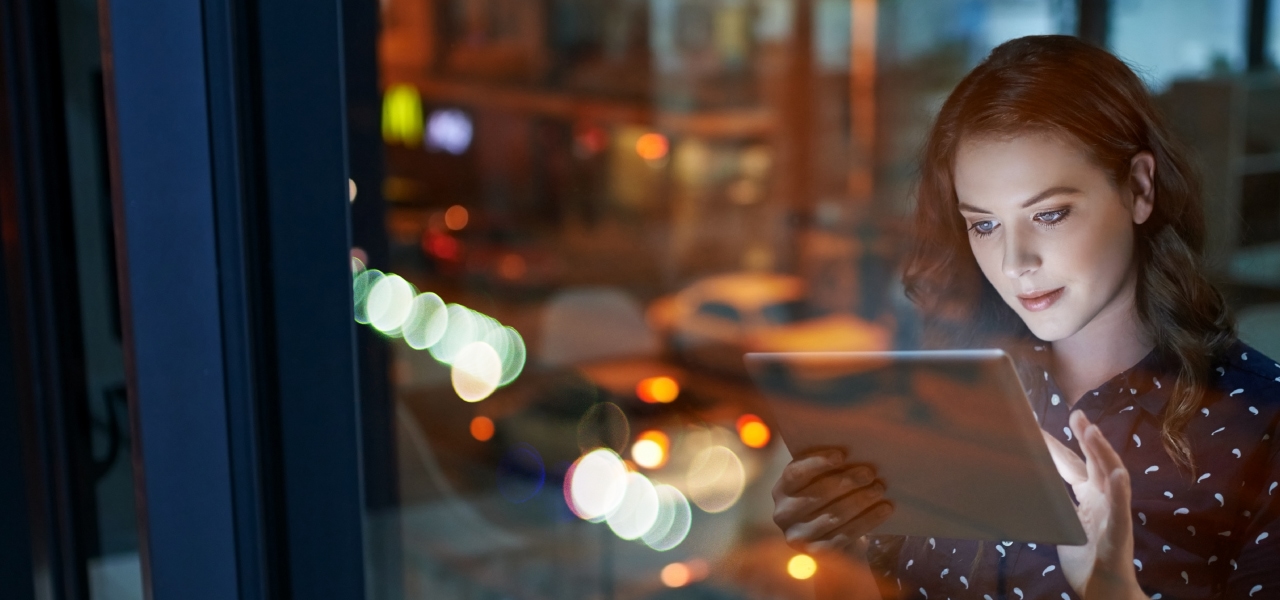 Woman using tablet by window at night. Blurred city lights visible outside. No readable text.