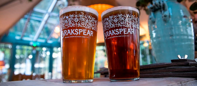 Two pint glasses of beer on a table. The glass on the left contains a lighter amber beer, and the one on the right holds a darker brown beer. Both glasses have decorative white patterns and the text 