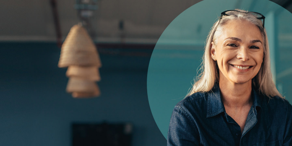 Smiling woman with long gray hair. She is wearing a denim shirt and glasses on her head. In the background, there is a blurred hanging light fixture and a blue wall. No visible text.