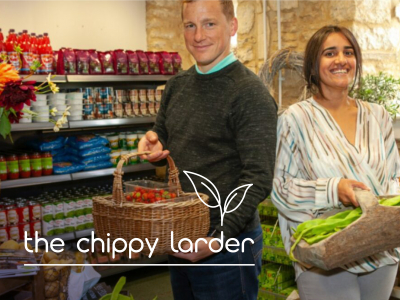 Two people holding baskets of produce. Shelves with various packaged goods in the background. Visible text: "the chippy larder" with a leaf design.