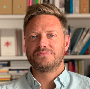 Man with short hair in front of bookshelf. Books and folders are visible in the background. No readable text is present.