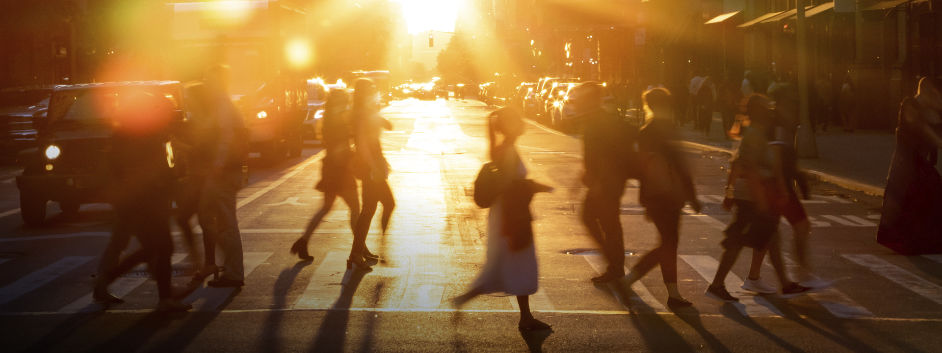 Street scene with people walking during sunset, with light flares from cars and a warm, glowing atmosphere.