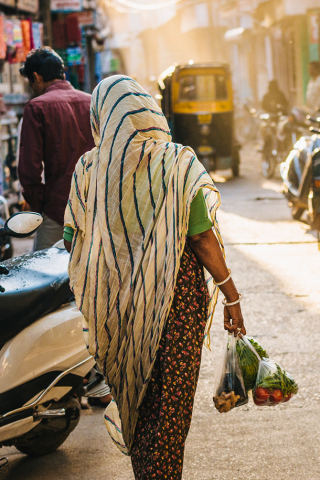Woman carrying bags of vegetables while walking down a vibrant street, with sunlight filtering through the scene.