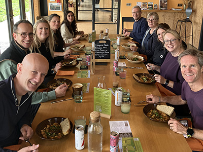 A group shot of the mark-making* team sitting at a long wooden table inside the FarmEd café, smiling at the camera while enjoying their meal together.